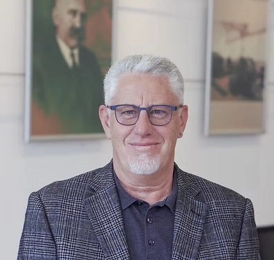 Herr Brantner, mit grauem Haar und Brille, in kariertem Sakko vor Bürohintergrund mit gerahmten Bildern bei der Fernwärme Wattens GmbH