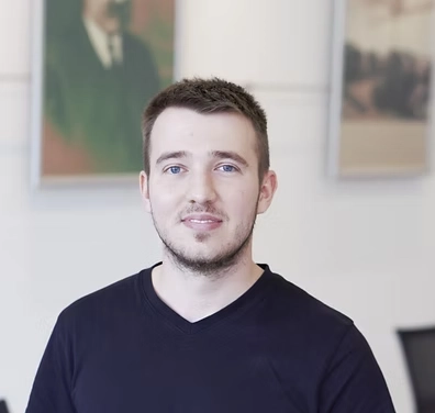 Porträt von Herrn Erlacher. Er trägt ein dunkelblaues T-Shirt und blickt mit einem freundlichen Lächeln direkt in die Kamera. Er hat kurze, dunkle Haare und blaue Augen. Der Hintergrund ist hell und zeigt zwei unscharfe gerahmte Bilder an einer Wand.