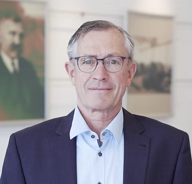 Porträt von Herrn Haim der Fernwärme Wattens. Er trägt ein dunkelblaues Sakko über einem hellblauen Hemd und eine Brille mit dunklem Gestell. Er blickt freundlich direkt in die Kamera. Der Hintergrund ist hell und zeigt unscharf angedeutete Bilder an einer Wand.