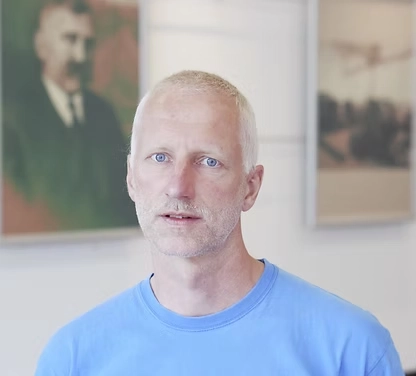 Porträt von Herrn Magerl. Er trägt ein hellblaues T-Shirt und blickt direkt in die Kamera. Er hat kurze, helle Haare und blaue Augen. Der Hintergrund ist hell und zeigt zwei unscharf hängende Bilder an einer Wand.