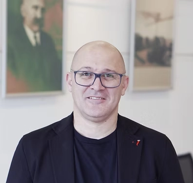 Porträt von Herrn Reinmayr. Er trägt ein schwarzes Sakko über einem schwarzen T-Shirt und eine blaue Brille. Er lächelt freundlich direkt in die Kamera. Der Hintergrund ist hell und zeigt unscharf hängende Bilder an einer Wand.