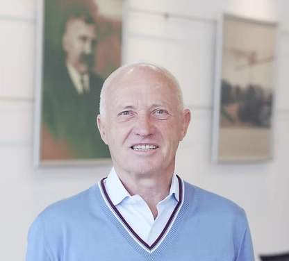 Herr Schmadl, lächelnd, in hellblauem Pullover vor neutralem Hintergrund mit gerahmten Bildern im Büro der Fernwärme Wattens GmbH